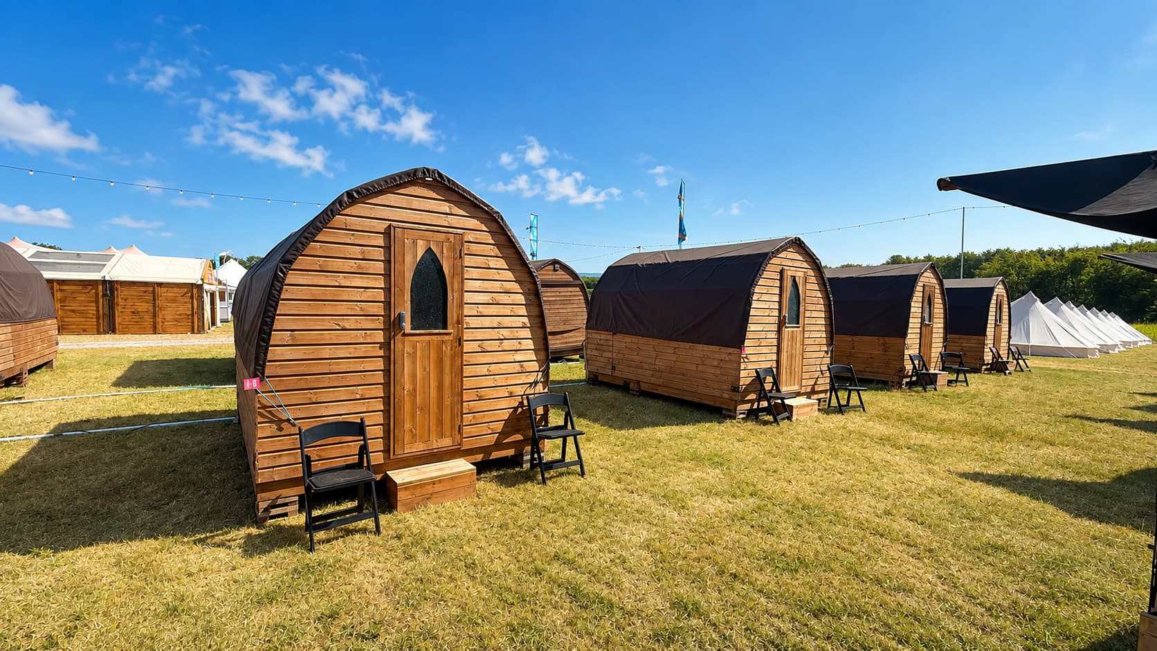 A row of wooden camping pods with dark roofs at Hotel Melody's Private Lodge, set on grass under a clear blue sky, with bell tents visible beyond.