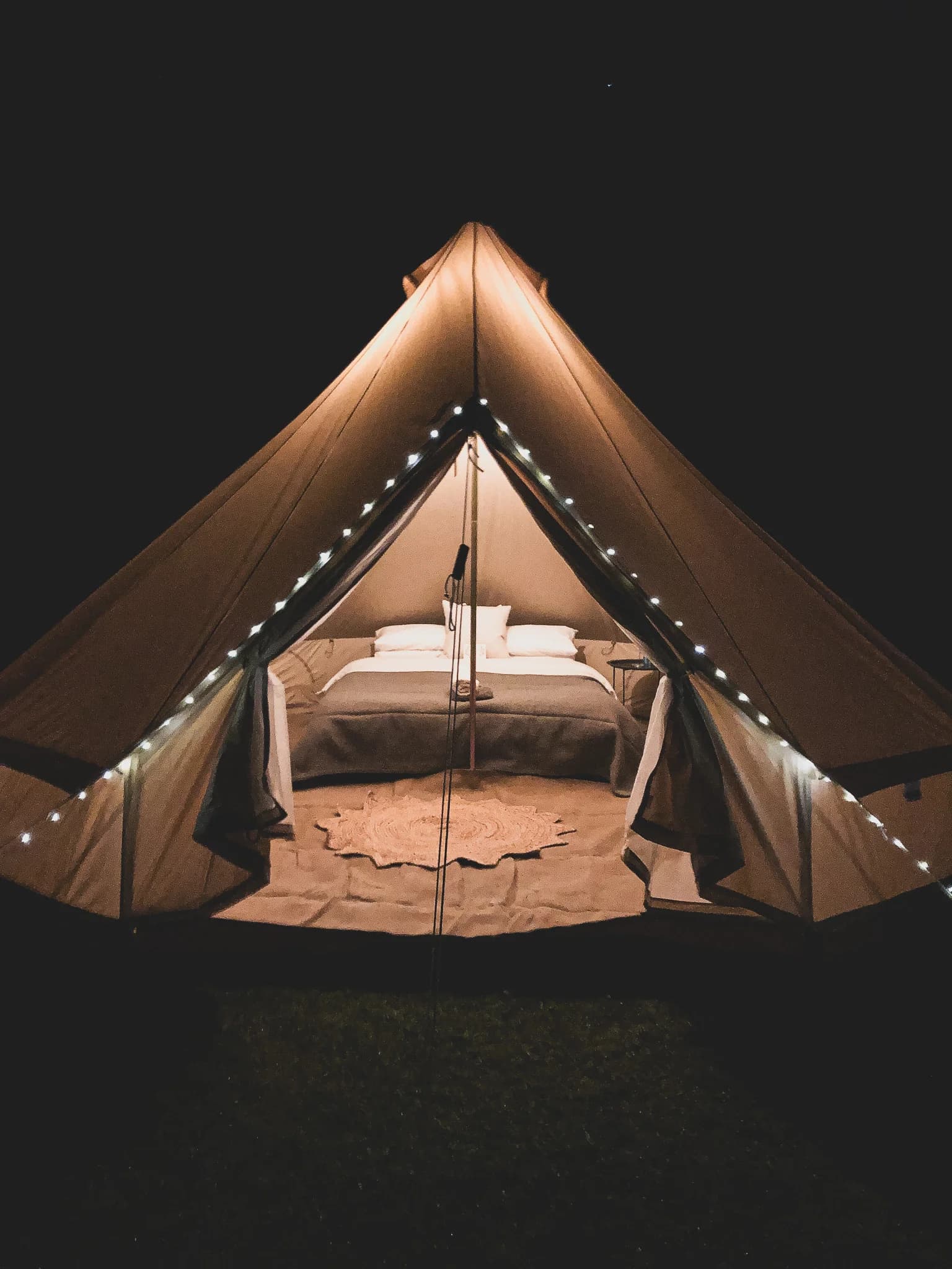 A Hotel Melody Lyrical Lounge bell tent lit up at night, with fairy lights along its seams and a made-up bed visible inside.