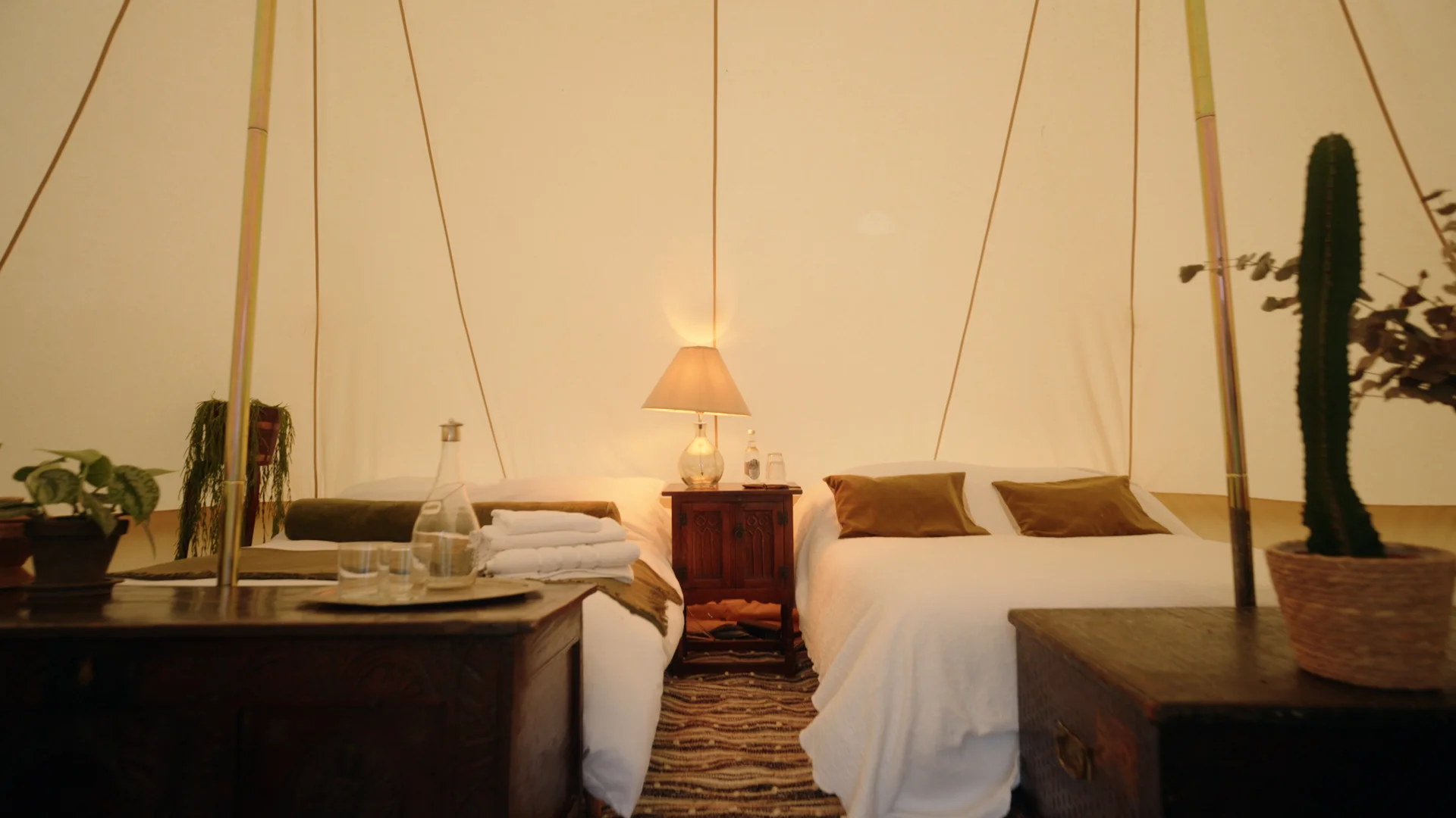 Twin beds dressed with white linen and mustard cushions inside a Hotel Melody Rhythm Resort tent, with potted plants and a cactus.