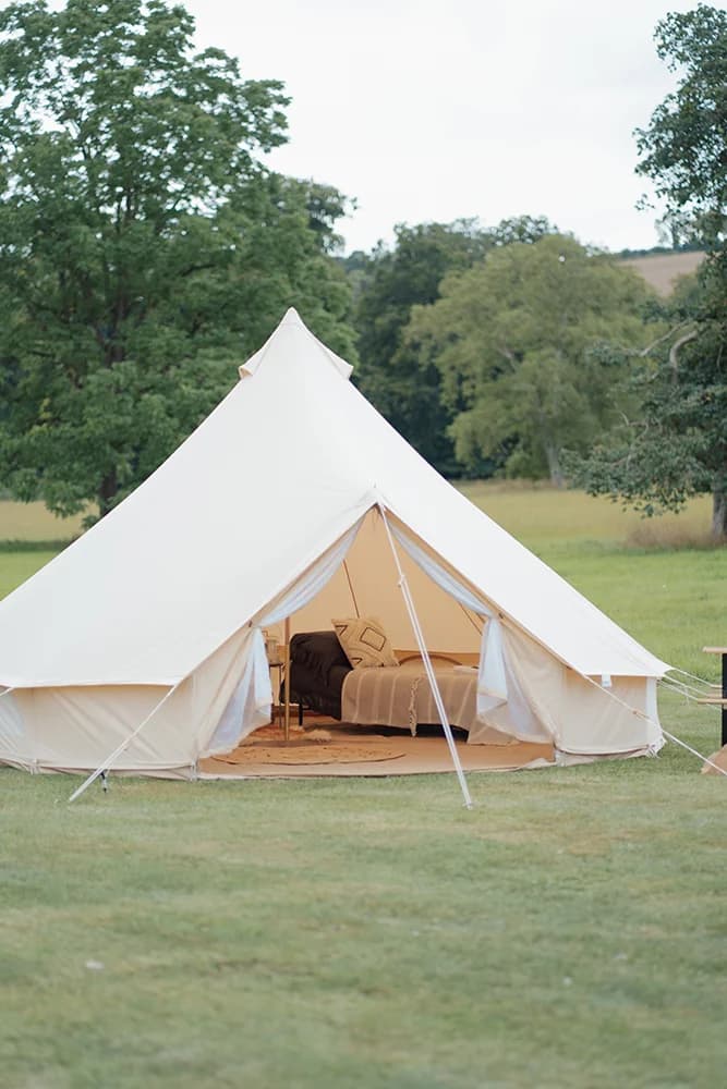 The exterior of a white bell tent at Hotel Melody's Lyrical Lounge, its open entrance revealing a dressed bed inside, set in a grassy field.