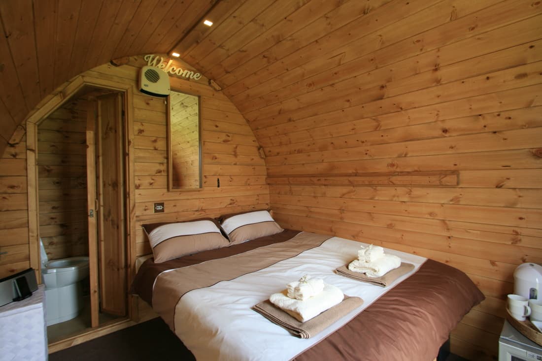 Interior of a wooden camping pod at Hotel Melody's Private Lodge, with a made-up double bed, folded towels, and an en-suite doorway visible.